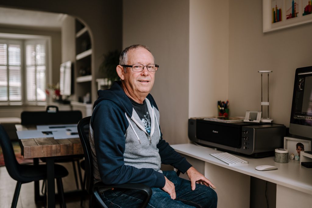 Man in dark and light grey top sitting on a office chair. In front is a desk with a computer and printer.