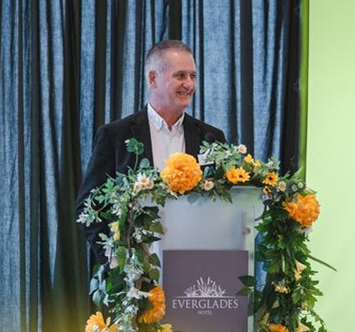 Man standing behind a podium. Podium has flowers around it and a logo with the name Everglades.