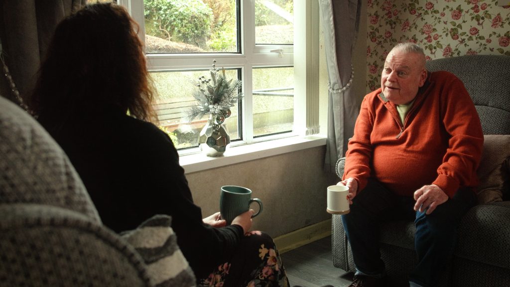 Man in orange jumper sitting on chair holding a mug in his right hand. Talking to a woman sitting opposite him.